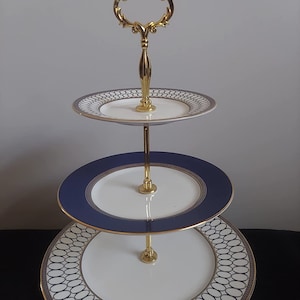 May include: Three-tiered cake stand with a gold metal handle and base. The stand has white porcelain plates with a blue and gold geometric pattern.