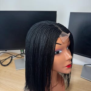 May include: A black braided wig with a blunt cut, displayed on a mannequin head. The wig has a lace front and is styled on a mannequin with red lipstick. The background features two black computer monitors.