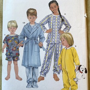 May include: Simplicity sewing pattern number 5874 for children's pyjamas. The pattern includes four different styles: a blue robe with a white stripe, a striped pyjama set, a yellow one-piece pyjama, and a pyjama top with a playful print. The pattern is for sizes AA 1/2, 1, and 2.