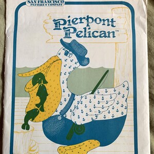 Puede incluir: Un patrón de costura vintage para un pelícano de 56 cm con una caña de pescar. El patrón se titula "Pierpont Pelican" y presenta un pelícano de dibujos animados con un sombrero azul y un mono. El pelícano está sentado en un muelle de madera con un pez en su pico.