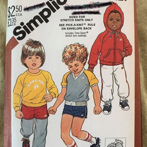 May include: Simplicity sewing pattern number 5591 for children's clothing. The pattern is for stretch knits only and includes three different designs: a yellow sweatshirt with red shorts, a blue and grey shirt with blue shorts, and a red hooded sweatshirt with white pants. The pattern includes blue wax transfers for embroidery or paint.