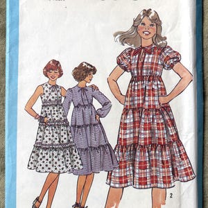 May include: Simplicity 8504 sewing pattern showing three tiered dresses. The dresses are in different styles and patterns, including a red and white plaid, a floral print, and a solid purple. The pattern is for sizes 5/6, YNG. JR./TEEN.