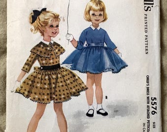 May include: Vintage McCall's printed pattern for a child's dress with an attached petticoat. The cover features illustrations of two young girls wearing the dresses. The dress on the left is brown with a floral pattern, and the dress on the right is blue.