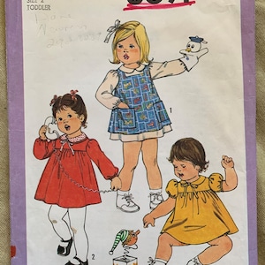 May include: Simplicity sewing pattern number 8691 for toddler girls' dresses. The pattern includes three different dress styles: a red dress with a white collar, a yellow dress with a white collar, and a blue and white patterned dress with a white collar. The pattern also includes instructions for making a toy puppet.