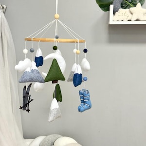 Könnte beinhalten: Ein hängendes Mobile für das Kinderzimmer mit Filzfiguren an einem runden Holzrahmen. Die Elemente umfassen graue und weiße Berge, grüne Tannenbäume, einen weißen Halbmond, weiße Wolken, dunkelgraue Skier sowie hellblaue Skischuhe aus Filz.