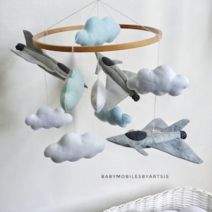 Puede incluir: Un móvil colgante con tres aviones de combate de fieltro gris y varias nubes suaves en tonos blanco, azul claro y verde pálido suspendidos de un marco circular de madera. El texto BABYMOBILESBYARTSIS está impreso en negro en la parte inferior.