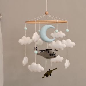 May include: A hanging mobile featuring a circular wooden frame with white strings suspending a light blue crescent moon, several white clouds, a grey helicopter, a green airplane, and small light green felt balls arranged in a layered design.