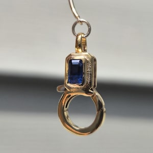 Puede incluir: Un cierre de mosquetón de oro con un zafiro azul de talla esmeralda rectangular engastado en la parte superior. El cierre incluye un mecanismo de anillo circular en la base y una pequeña anilla en la parte superior para su fijación.