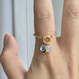 May include: A gold ring with a delicate chain band and a circular charm holding three gemstone pendants. The pendants include a blue, a white, and a blue-and-white stone. The ring is on a finger.