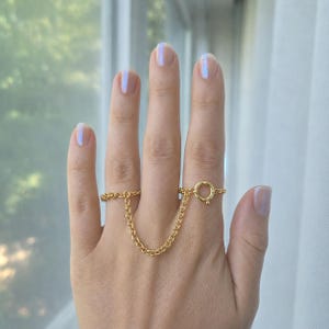 May include: A gold-toned double ring connected by a delicate chain. The piece features a circular spring ring clasp mechanism on one side, linking two separate bands that sit on adjacent fingers, creating a continuous linked jewelry design.