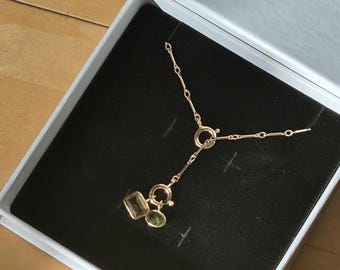 May include: A gold-toned chain necklace featuring a Y-shaped design with a central circular clasp. Two faceted gemstones, one rectangular and light yellow and one oval and green, are suspended from the clasp as decorative charms.