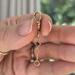 May include: A gold watch chain fob featuring a swivel clasp at the top, a small diamond-shaped link set with a dark brown gemstone, and a vertical bar connector ending in a circular loop at the bottom, all crafted from polished yellow gold metal.