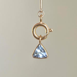 Puede incluir: Un cierre de resorte en tono dorado unido a una barra de metal vertical, con un pequeño dije de piedra preciosa triangular de color azul claro facetado suspendido de la parte inferior del mecanismo de cierre circular, creando un componente de joyería.