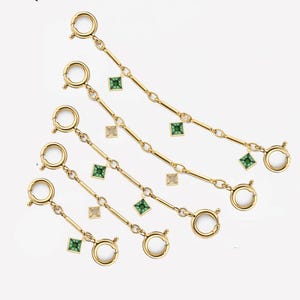 May include: Gold-toned chain extenders with spring ring clasps and small square gemstones in green and champagne colors. The extenders are arranged diagonally against a white background.