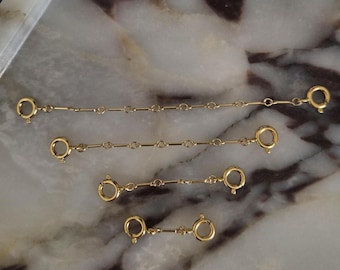 May include: Four gold-toned metal jewelry extender chains of varying lengths, each featuring a circular spring ring clasp at both ends. The chains are arranged horizontally in descending order of length, showcasing a simple link design.