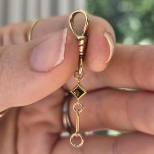 May include: A gold-toned metal watch fob or pendant connector featuring a swivel clasp at the top, a small square-shaped charm with a dark brown gemstone inlay, and a short dangling chain ending in a circular jump ring.