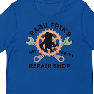 May include: A royal blue t-shirt featuring a black graphic of a repair shop logo. The logo depicts a silhouette of a person working on a gear with wrenches on either side. The text "Babu Frik's Repair Shop" is written in black with the words "Hey" and "Heeeyyy" on either side of the gear.