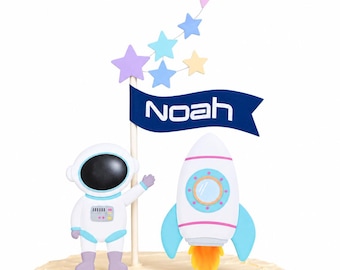 May include: A cake topper featuring a white astronaut figure, a white and blue rocket ship with orange flames, and a dark blue banner with the white text Noah. The topper includes a vertical stick adorned with several pastel-colored stars.