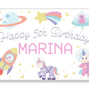 Op de afbeelding: Een kleurrijk verjaardagsbanner met een ruimtethema. Het bevat een raket, een eenhoorn, een planeet, een vallende ster en een UFO. De banner zegt "Happy 5th Birthday MARINA".