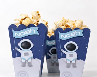 May include: Three dark blue popcorn containers featuring a white astronaut graphic on a gray lunar surface. Each container has a light blue banner with the name Noah printed in white text. The containers are filled with popped popcorn.