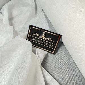 May include: White sheer fabric with a fine mesh texture. The fabric is folded and draped, showing its delicate and airy quality. A black card with gold lettering reads "Lovely Lace Couture".