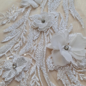 Peut inclure: Tissu de dentelle blanche avec une broderie florale délicate et de petits strass transparents. Le tissu présente un motif répétitif de feuilles et de fleurs, avec quelques fleurs tridimensionnelles plus grandes.