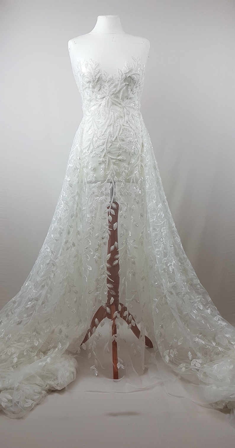 May include: White tulle fabric with a delicate floral pattern and silver sequins. The fabric is draped over a dress form.