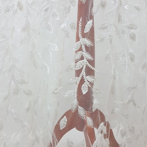 May include: White tulle fabric with a delicate leaf pattern embroidered with sequins. The fabric is sheer and has a soft, flowing texture.