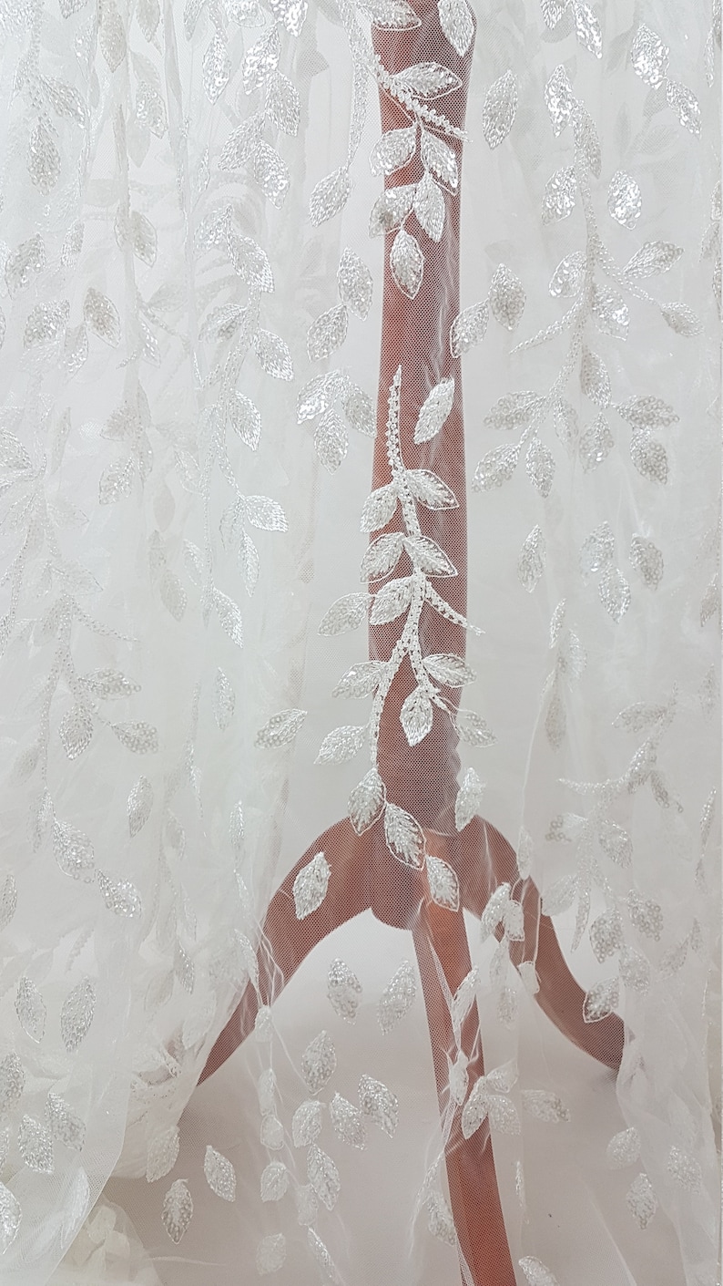 May include: White tulle fabric with a delicate leaf pattern embroidered with sequins. The fabric is sheer and has a soft, flowing texture.