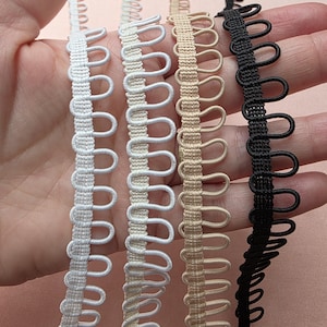 Op de afbeelding: Close-up van vier decoratieve sierbanden in verschillende kleuren: wit, crème, beige en zwart. Elke band heeft een lusvormig ontwerp langs één rand. De banden worden in een hand gehouden.