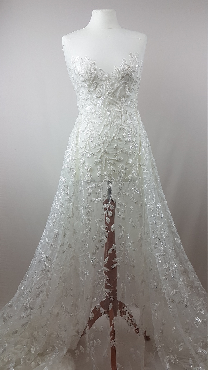 May include: White beaded lace fabric with a leaf design. The fabric is on a dress form.