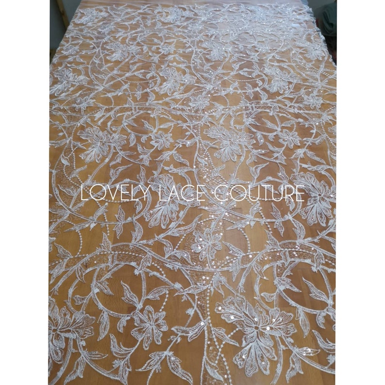 Może przedstawiać: Biała koronka kwiatowa z cekinami. Koronka ma delikatny wz&oacute;r kwiatowy z małymi cekinami rozrzuconymi po całej powierzchni. Koronka jest pokazana na drewnianej powierzchni. "LOVELY LACE COUTURE" jest nadrukowane na koronce.