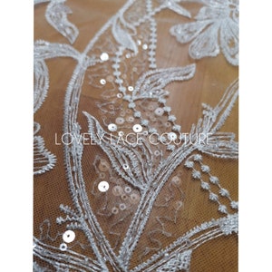 Może przedstawiać: Biała koronka kwiatowa z cekinami. Koronka ma delikatny wz&oacute;r kwiatowy z małymi białymi cekinami rozrzuconymi po całej powierzchni. Tekst "LOVELY LACE COUTURE" jest widoczny w środku obrazu.