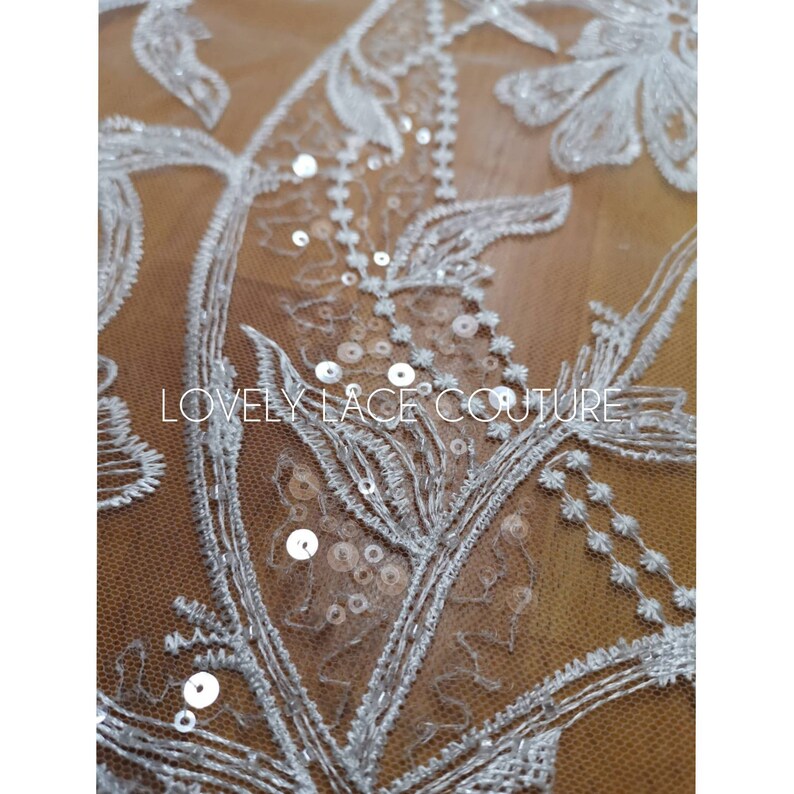 Może przedstawiać: Biała koronka kwiatowa z cekinami. Koronka ma delikatny wz&oacute;r kwiatowy z małymi białymi cekinami rozrzuconymi po całej powierzchni. Tekst "LOVELY LACE COUTURE" jest widoczny w środku obrazu.