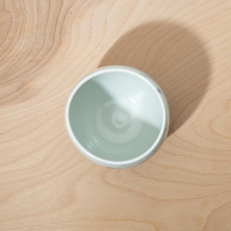 May include: A small, light green ceramic bowl with a rounded shape. The bowl is set against a light brown wooden surface, with a shadow cast to the side. The bowl's interior is a lighter shade of green.