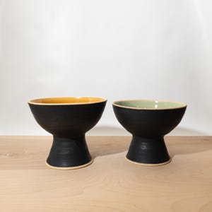 May include: Two ceramic pedestal bowls with matte black exterior surfaces and flared bases. The left bowl features a warm yellow interior glaze, while the right bowl has a soft sage green interior glaze. Both bowls have exposed clay rims.