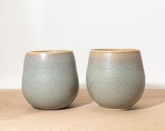 May include: Two ceramic stemless wine glasses with a speckled, muted blue-green glaze and a light brown rim. The rounded cups sit on a light wood surface. The glasses have a smooth, modern design.