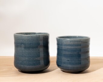 May include: Two handmade, blue ceramic cups with a textured surface. The cups have a cylindrical shape with a slightly wider base and a smooth rim. The blue color has variations, adding to the handcrafted aesthetic. The cups are suitable for beverages.
