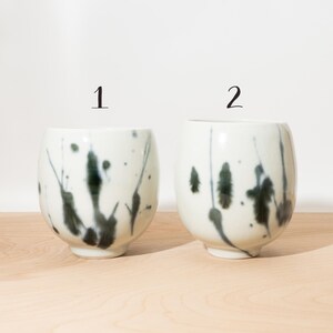May include: Two white ceramic cups with a speckled dark green and black design. The cups are numbered 1 and 2 in black. The cups have a rounded shape and sit on a light wood surface.