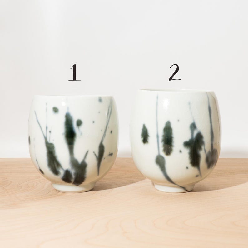 May include: Two white ceramic cups with a speckled dark green and black design. The cups are numbered 1 and 2 in black. The cups have a rounded shape and sit on a light wood surface.