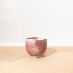 May include: A small, rounded ceramic cup featuring a mottled glaze in shades of dusty rose, mauve, and deep red. The surface displays a textured, crystalline pattern with subtle variations in color density, finished with a smooth, white rim.