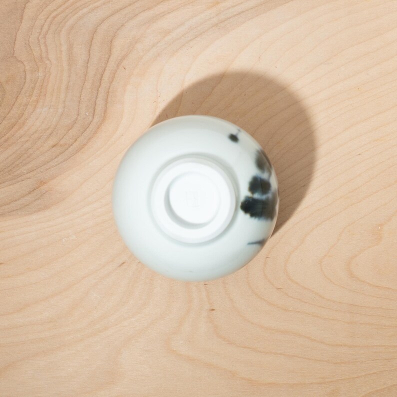 May include: A small, white ceramic bowl with a circular base and a slightly flared rim. The bowl features a dark, abstract pattern on the side. The bowl is set against a light-colored wooden surface.