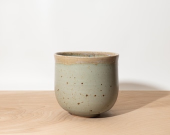 May include: A handmade ceramic cup with a rounded base and a light green glaze. The rim has a natural, unglazed tan color. The cup is speckled with small brown dots, giving it a rustic appearance. The cup is sitting on a wooden surface.