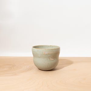 May include: A small, rounded ceramic cup with a speckled light green glaze. The vessel features a smooth, curved silhouette with subtle variations in color and texture across the surface, showcasing a hand-crafted appearance and a wide opening.