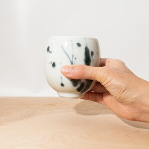 May include: A white ceramic cup with a rounded shape, featuring black ink splatter designs. The cup is held by a hand, showcasing the cup's form and the artistic ink pattern. The cup is on a light wood surface.