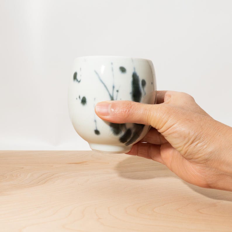 May include: A white ceramic cup with a rounded shape, featuring black ink splatter designs. The cup is held by a hand, showcasing the cup's form and the artistic ink pattern. The cup is on a light wood surface.