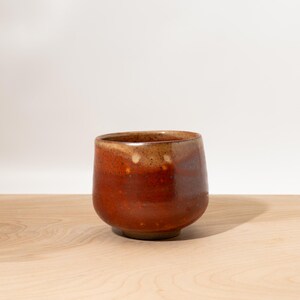 May include: A handmade ceramic cup with a rounded base and a wide opening. The cup features a rich, earthy color palette, with shades of brown and reddish-orange. The interior and rim have a lighter, tan hue. The cup sits on a wooden surface.
