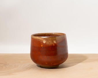May include: A handmade ceramic cup with a rounded base and a wide opening. The cup features a rich, earthy color palette, with shades of brown and reddish-orange. The interior and rim have a lighter, tan hue. The cup sits on a wooden surface.