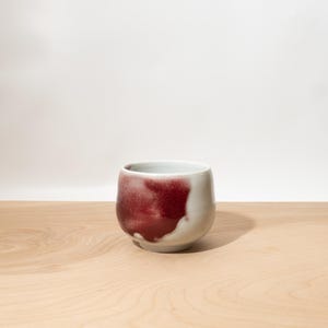 May include: A small, rounded ceramic cup featuring a smooth, off-white base glaze. One side of the exterior is decorated with a prominent, textured splash of deep crimson red glaze that fades into the lighter background, creating a distinct contrast.