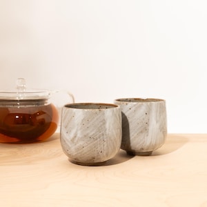 May include: Two speckled gray ceramic tea cups with a textured, swirled design. The cups have a rounded shape and a brown rim. A glass teapot filled with tea is visible in the background, suggesting a tea set.